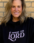 Woman wearing a black hoodie with 'Praise the Lord' text against a brick wall.