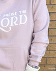 Light pink hoodie with 'Praise the Lord' text against a brick wall.