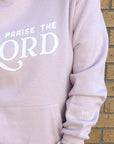 Light pink hoodie with 'Praise the Lord' text against a brick wall.
