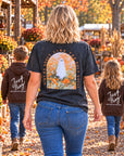 A woman and two children walk down a fall-themed pumpkin patch aisle, viewed from behind. The woman wears a dark charcoal graphic T-shirt with a ghost illustration and the words 'Ain't no ghost like the holy Ghosts,' while the children wear matching brown hoodies with 'Just Pray' written on the back.