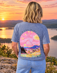 A woman stands on a rocky overlook at sunset with a lake and forested hills in the background, wearing a blue-gray graphic T-shirt from the back. The shirt features a large colorful print inspired by Van Gogh, with swirling pink and yellow sky, a village, and the text 'THE STARS WERE MADE TO WORSHIP SO WILL A'.