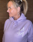 Woman wearing a lavender hoodie with 'Just Pray' text against a brick wall.