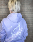 Person wearing a light blue hoodie with 'Just Pray' text on the back against a brick wall.