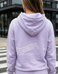 Person wearing a lavender hoodie with text on the back, standing outdoors.