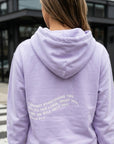 Person wearing a lavender hoodie with text on the back, standing outdoors.