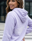 Woman wearing a lavender hoodie with text on the back, standing outdoors.