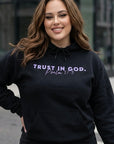 Woman wearing a black hoodie with 'Trust in God' text, standing outdoors.