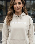 Woman wearing a beige hoodie with 'Trust in God' text, standing outdoors.