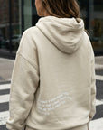 Person wearing a beige hoodie with text on the back, standing on a street.