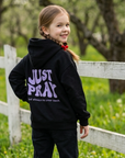 A young girl standing outdoors in a grassy field with a white fence and trees in the background, wearing a black hoodie with purple text reading 'JUST PRAY' on the back and black pants.