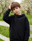 A young boy wearing a black pullover hoodie and matching black sweatpants stands in a grassy field with a white wooden fence and blooming trees in the background.