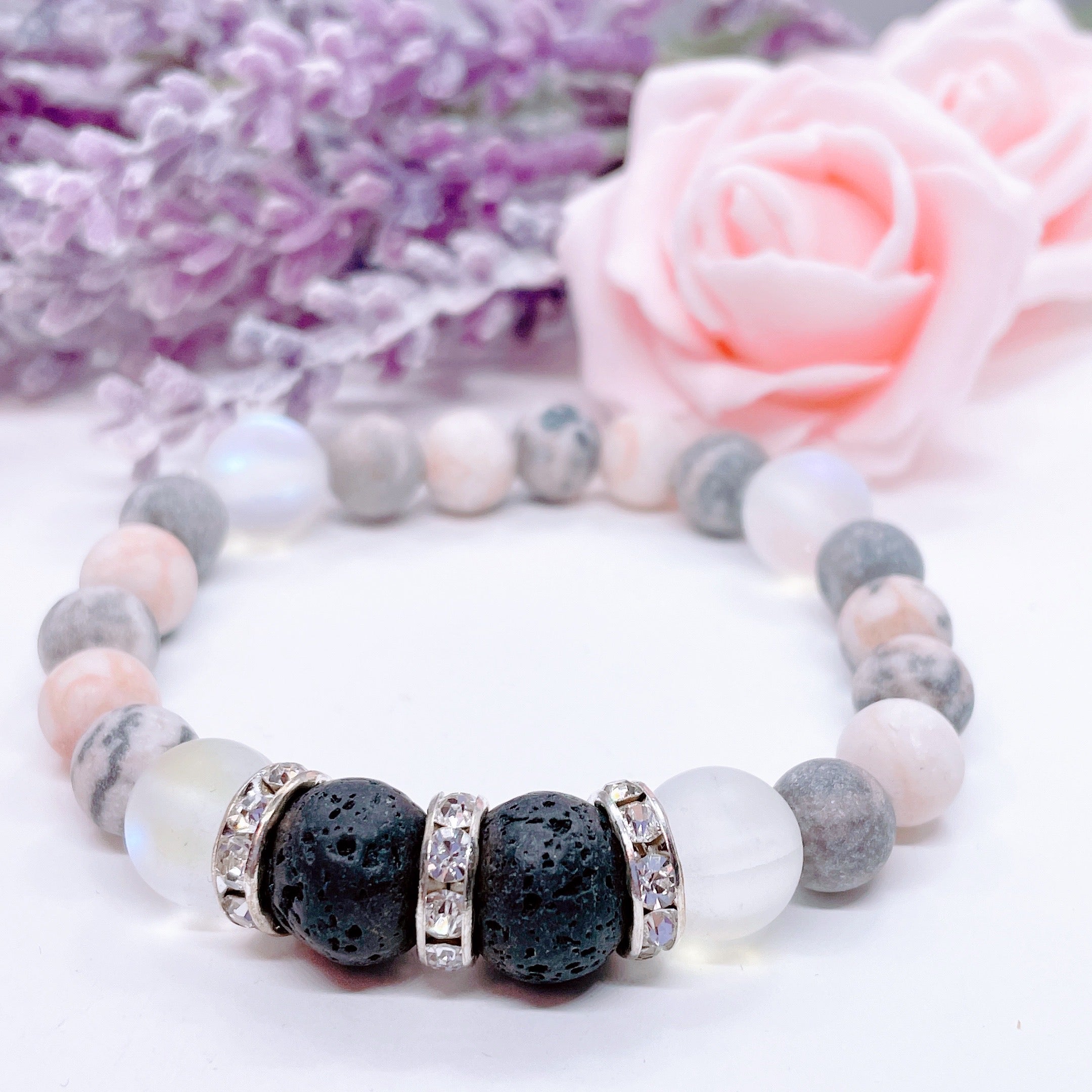 A Lava Stone and Rhinestone stretch bracelet made with 2 rough black lava stones, translucent aura beads, pink zebra jasper gemstones, and rhinestone accents for added sparkle sits on a white table. 