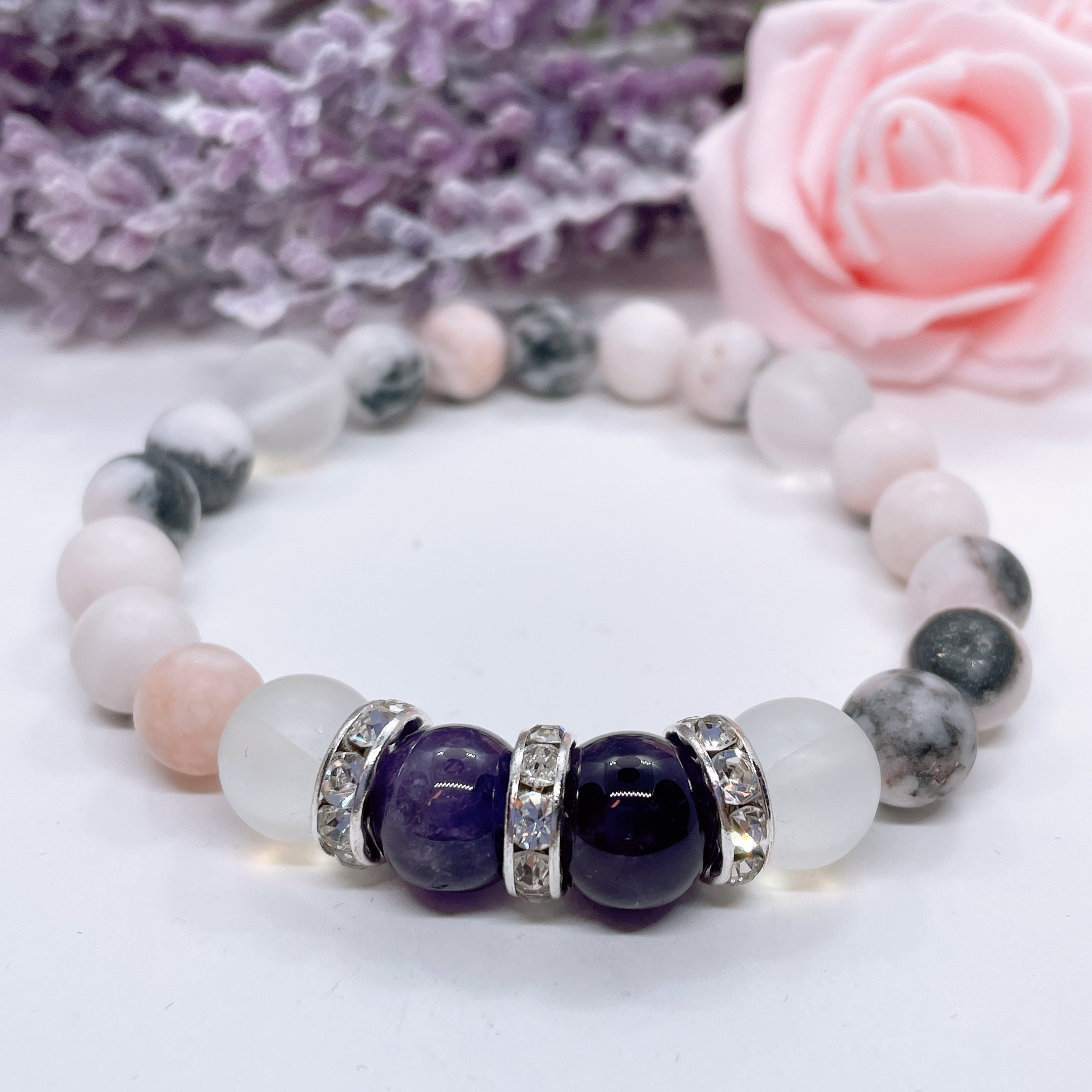An Amethyst Rhinestone and Gemstone stretch bracelet made with 2 dark purple amethyst gemstones, translucent aura beads, and rhinestone accents for added sparkle sits on a white table. 