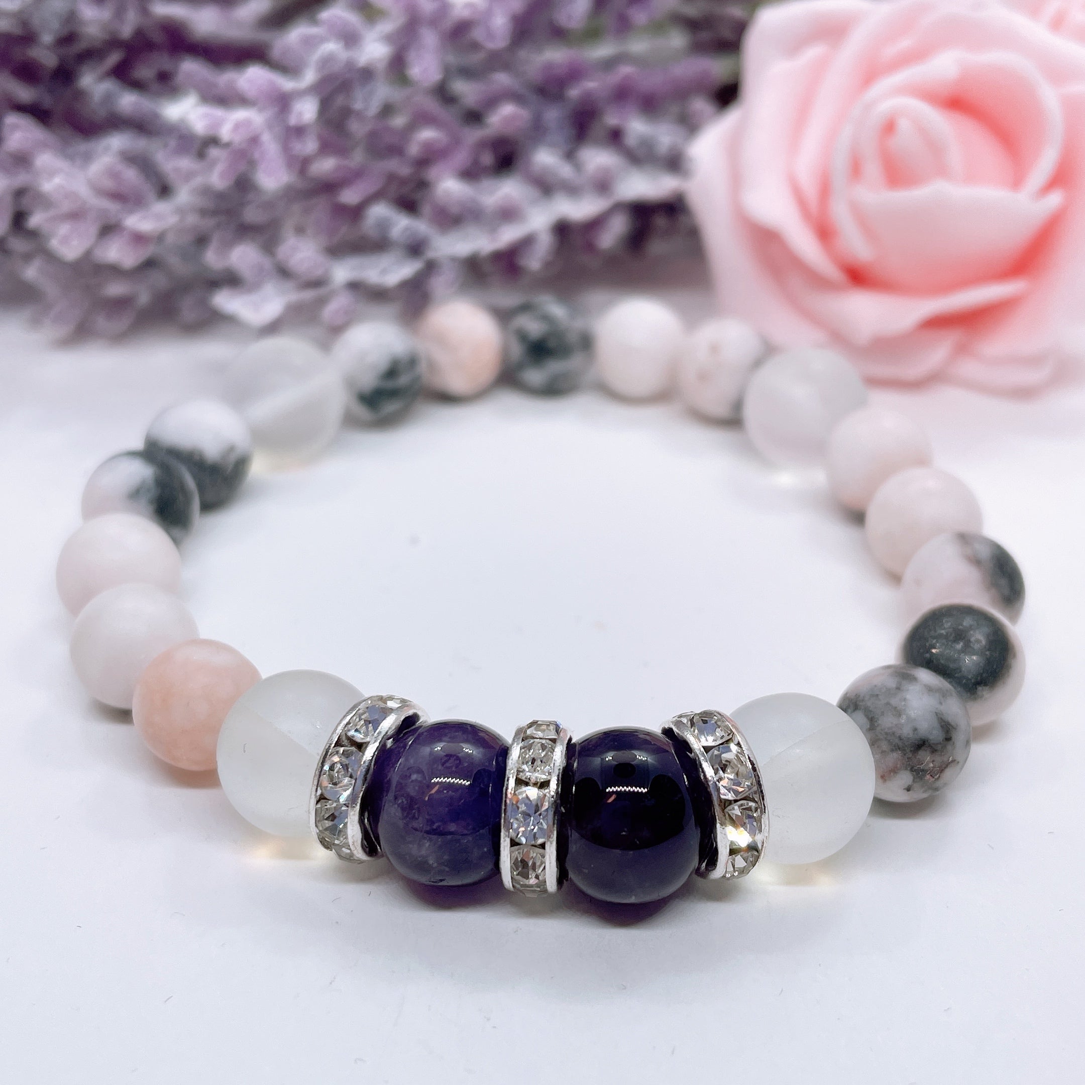 An Amethyst Rhinestone and Gemstone stretch bracelet made with 2 dark purple amethyst gemstones, translucent aura beads, and rhinestone accents for added sparkle sits on a white table.