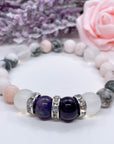 An Amethyst Rhinestone and Gemstone stretch bracelet made with 2 dark purple amethyst gemstones, translucent aura beads, and rhinestone accents for added sparkle sits on a white table.