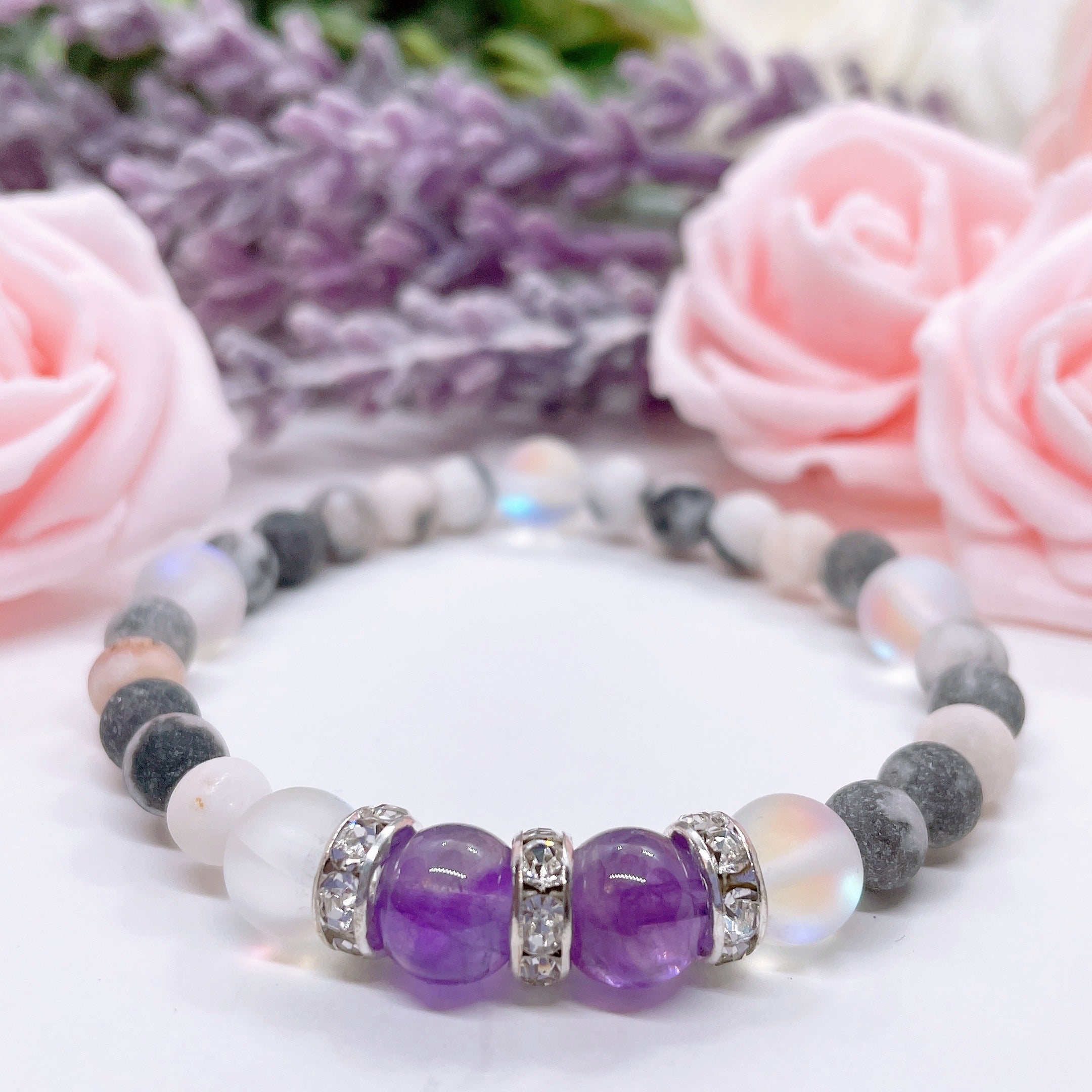 The Amethyst Rhinestone Companion Gemstone stretch bracelet made with 2 dark purple amethyst gemstones, translucent aura beads, and rhinestone accents for added sparkle sits on a white table.