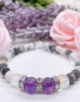 The Amethyst Rhinestone Companion Gemstone stretch bracelet made with 2 dark purple amethyst gemstones, translucent aura beads, and rhinestone accents for added sparkle sits on a white table.