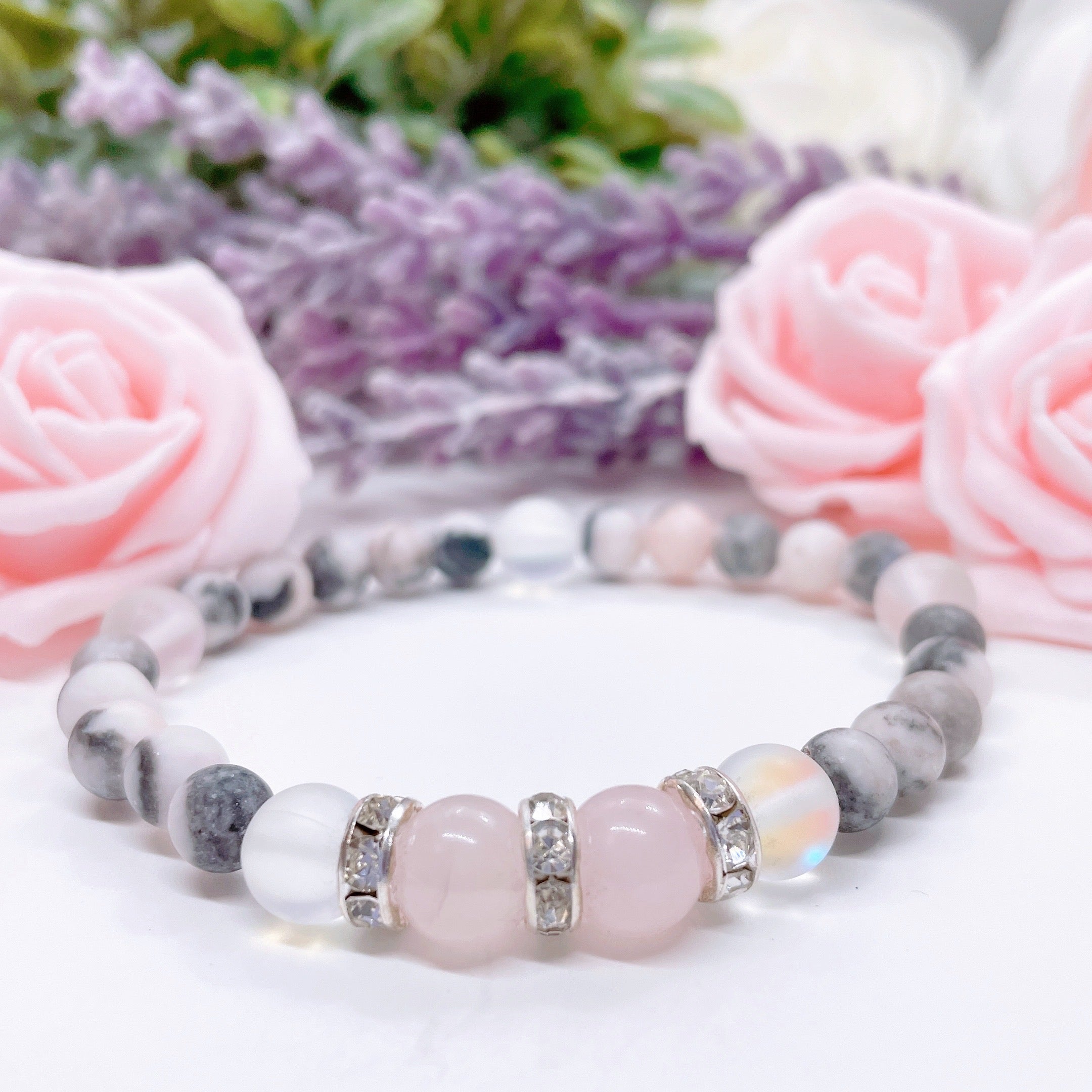 A Rose Quartz Rhinestone Companion Gemstone stretch bracelet made with 2  pink rose quartz gemstones, translucent aura beads, pink jasper gemstones, and rhinestone accents for added sparkle sits on a white table.