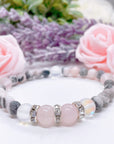 A Rose Quartz Rhinestone Companion Gemstone stretch bracelet made with 2  pink rose quartz gemstones, translucent aura beads, pink jasper gemstones, and rhinestone accents for added sparkle sits on a white table.