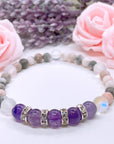 The Amethyst Rhinestone Companion Gemstone stretch bracelet made with 5 dark purple amethyst gemstones, translucent aura beads, and rhinestone accents for added sparkle sits on a white table.