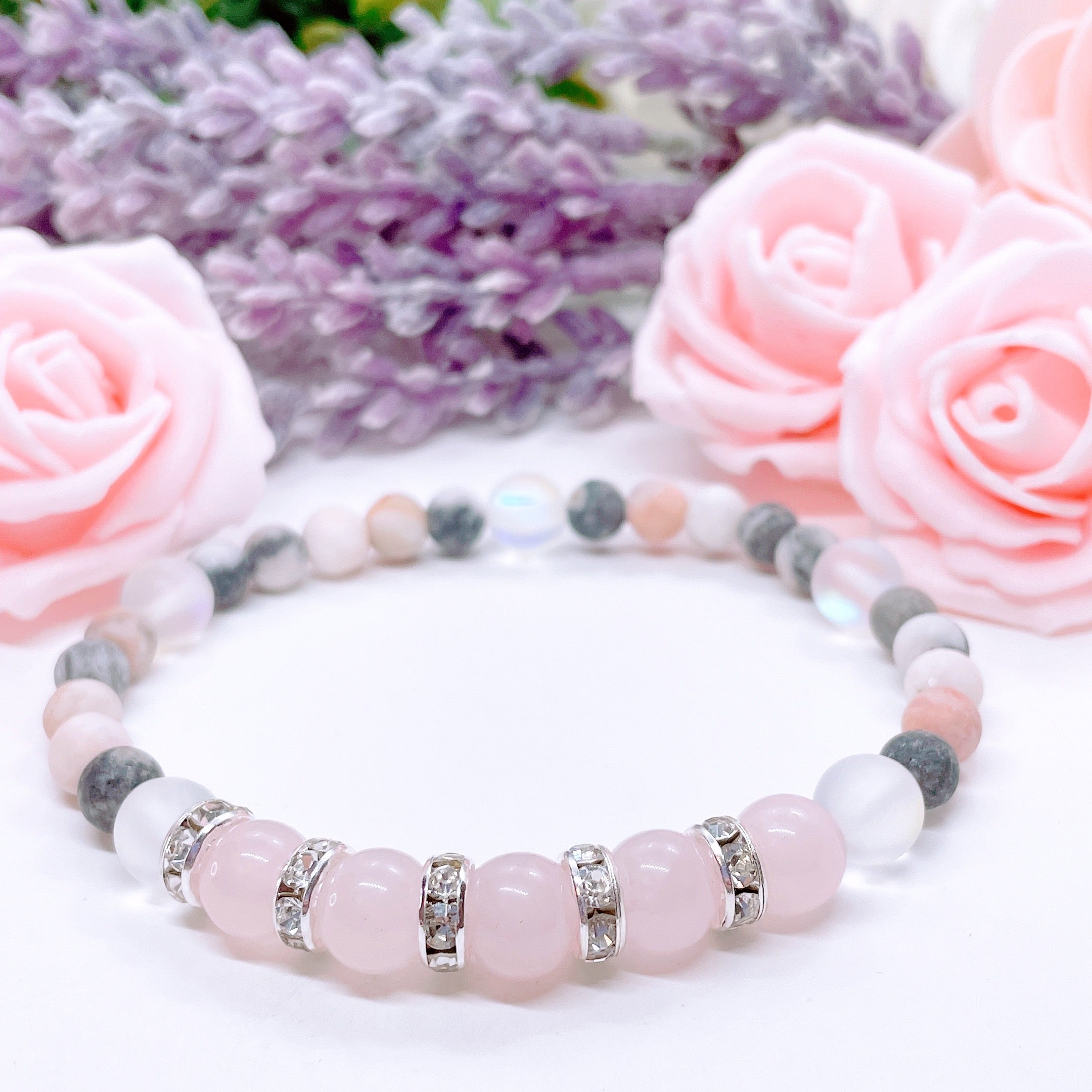 A Rose Quartz Rhinestone Companion Gemstone stretch bracelet made with 5 pink rose quartz gemstones, translucent aura beads, pink jasper gemstones, and rhinestone accents for added sparkle sits on a white table.