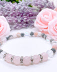 A Rose Quartz Rhinestone Companion Gemstone stretch bracelet made with 5 pink rose quartz gemstones, translucent aura beads, pink jasper gemstones, and rhinestone accents for added sparkle sits on a white table.