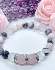A Rose Quartz Rhinestone and Gemstone stretch bracelet made with 2  pink rose quartz gemstones, translucent aura beads, pink jasper gemstones, and rhinestone accents for added sparkle sits on a white table.