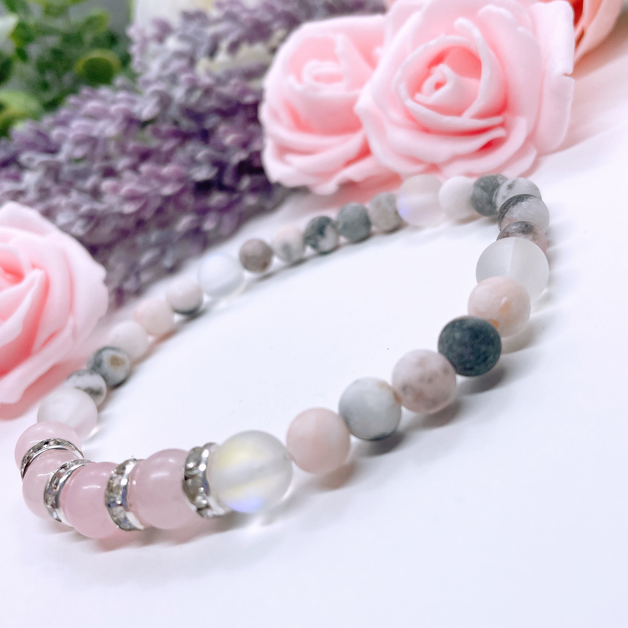 A Rose Quartz Rhinestone Companion Gemstone stretch bracelet made with 4 pink rose quartz gemstones, translucent aura beads, pink jasper gemstones, and rhinestone accents for added sparkle sits on a white table.