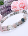 A Rose Quartz Rhinestone Companion Gemstone stretch bracelet made with 4 pink rose quartz gemstones, translucent aura beads, pink jasper gemstones, and rhinestone accents for added sparkle sits on a white table.