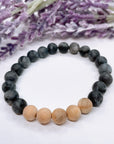 A black beaded bracelet with 18 black labradorite beads with 5 cedar wood brown beads on a white table with a purple flower on the table.