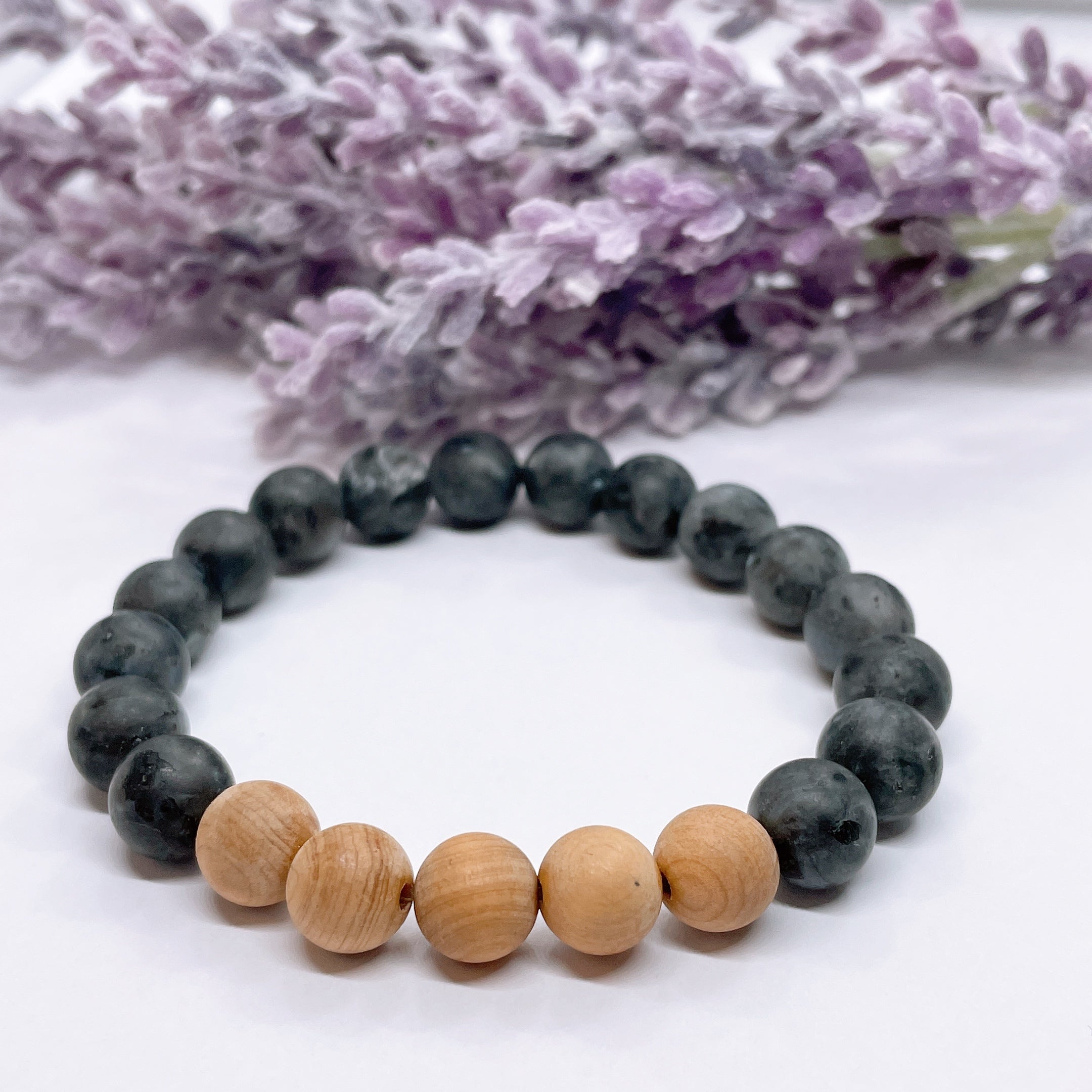 A black beaded bracelet with 16 black labradorite beads with 5 cedar wood brown beads on a white table with a purple flower on the table.