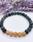 A black beaded bracelet with 22 black labradorite beads with 5 cedar wood brown beads on a white table with a purple flower on the table.