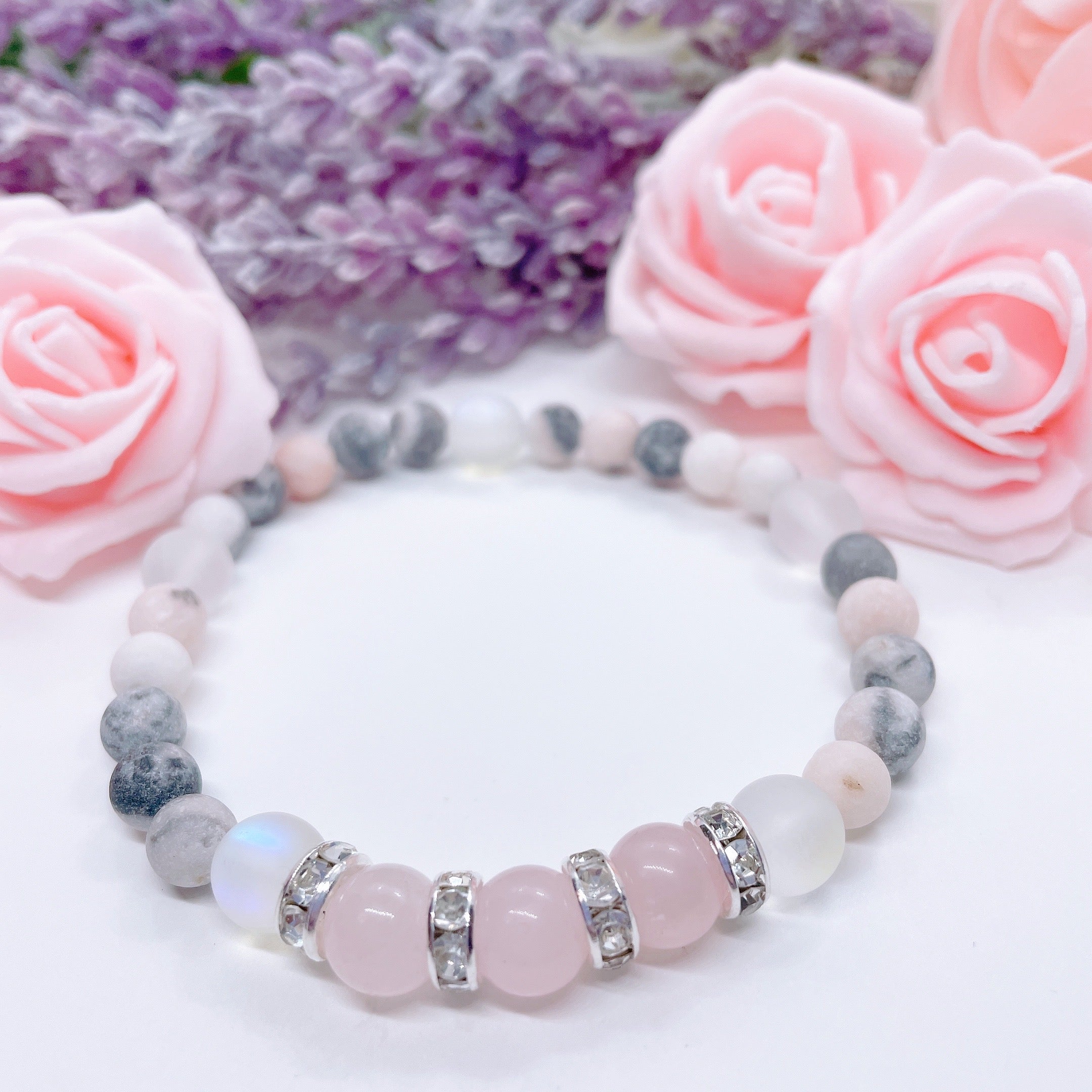 A Rose Quartz Rhinestone Companion Gemstone stretch bracelet made with 3 pink rose quartz gemstones, translucent aura beads, pink jasper gemstones, and rhinestone accents for added sparkle sits on a white table.