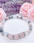 A Rose Quartz Rhinestone Companion Gemstone stretch bracelet made with 3 pink rose quartz gemstones, translucent aura beads, pink jasper gemstones, and rhinestone accents for added sparkle sits on a white table.