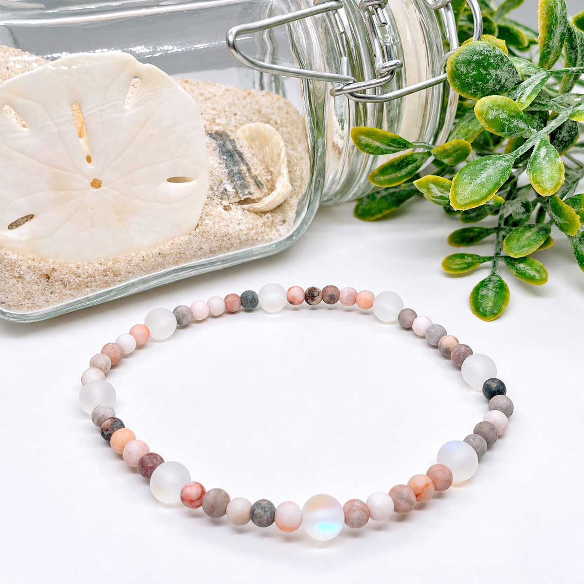 A beaded stretch bracelet made of round stone beads in soft pink, peach, gray, white, and iridescent opal-like tones, arranged on a white surface with decorative sand, a glass jar, and greenery in the background.