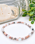 A beaded stretch bracelet made of round stone beads in soft pink, peach, gray, white, and iridescent opal-like tones, arranged on a white surface with decorative sand, a glass jar, and greenery in the background.
