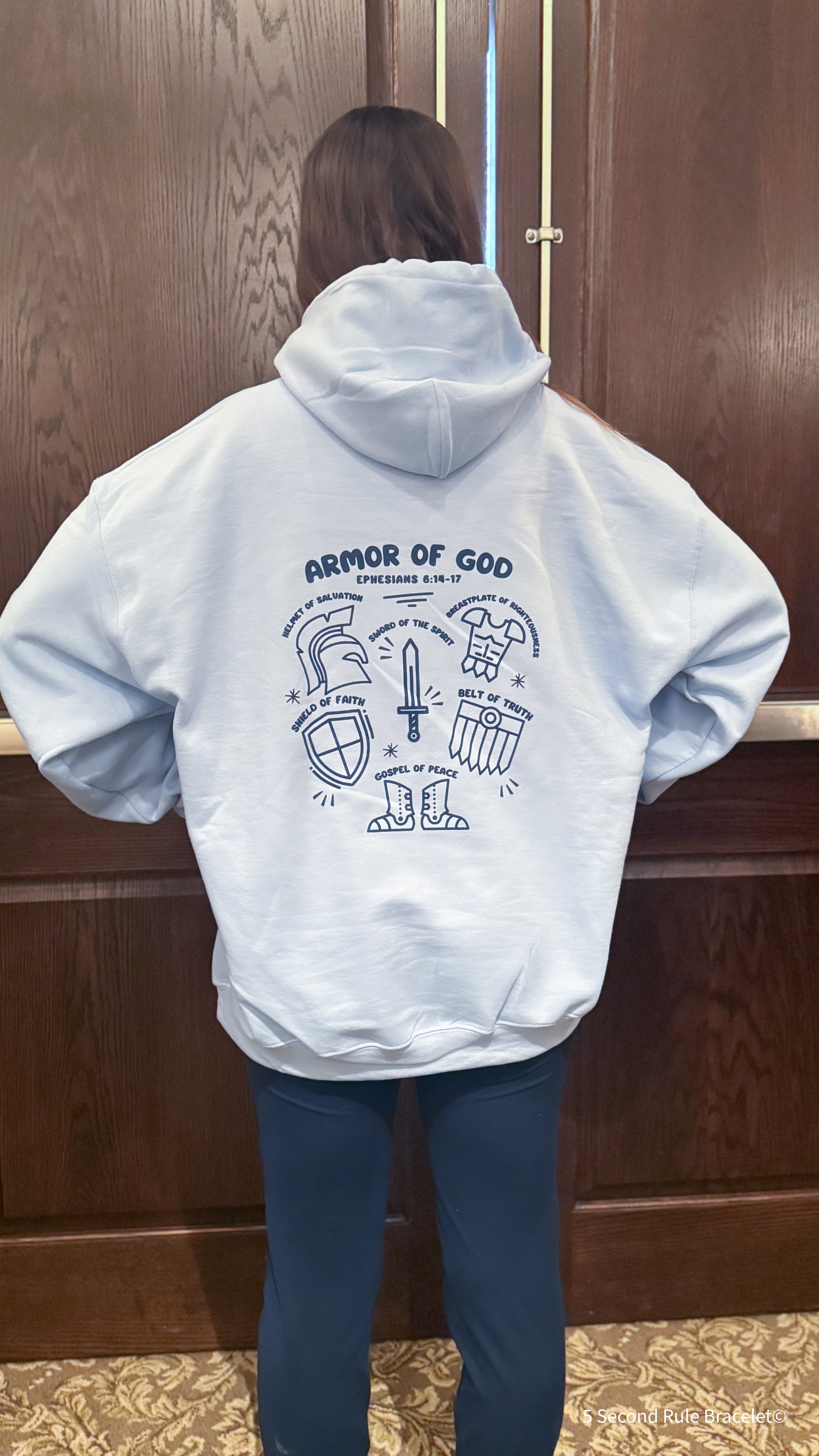 Person wearing a light blue hoodie with 'Armor of God' design on the back, standing against a wooden door.
