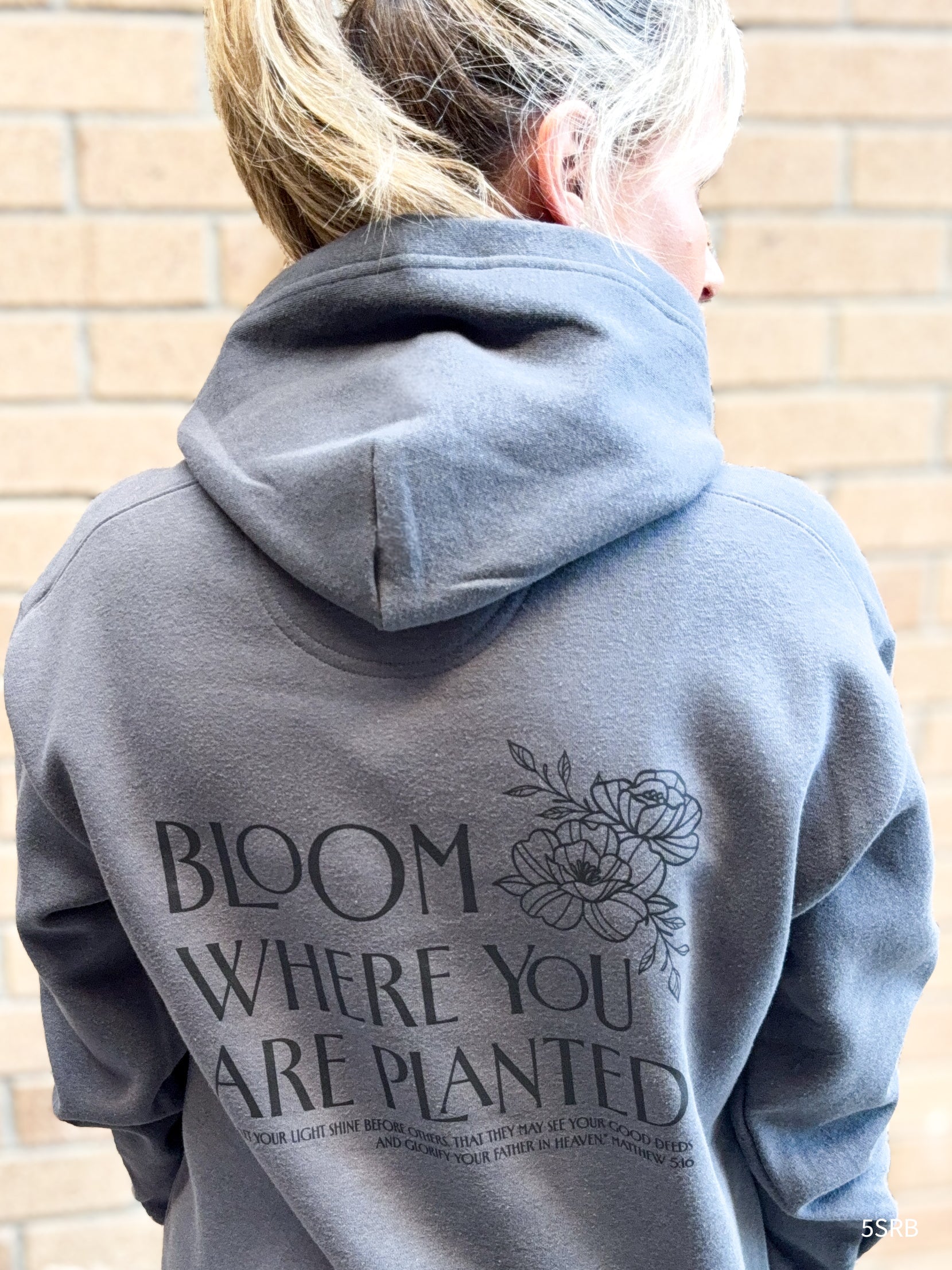 Person wearing a gray hoodie with text and floral design on the back, standing against a brick wall.