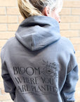 Person wearing a gray hoodie with text and floral design on the back, standing against a brick wall.