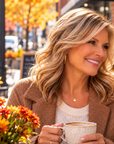 A women smiling and holding mugs at an outdoor patio table, with autumn trees, flowers, and a small pumpkin in the background.