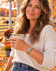 A woman in a cozy cream knit sweater and blue jeans stands outdoors on a porch holding a ceramic mug. She is wearing a gold-toned beaded bracelet with a silver-toned cross charm.