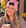 A smiling woman sitting outdoors at a cafe table, wearing a pink beanie with a black patch that reads 'Chosen Feminist' and holding a paper coffee cup.