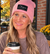 A smiling woman sitting outdoors at a cafe table, wearing a pink beanie with a black patch that reads 'Chosen Feminist' and holding a paper coffee cup.