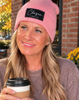 A smiling woman sitting outdoors at a cafe table, wearing a pink beanie with a black patch that reads 'Chosen Feminist' and holding a paper coffee cup.
