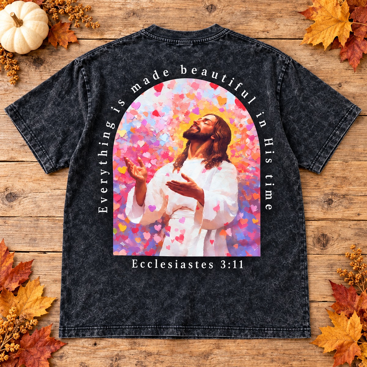 A black acid-wash graphic t-shirt laid flat on a rustic wooden background decorated with autumn leaves, a small pumpkin, and dried flowers. The back of the shirt features a large colorful graphic of Jesus in a white robe surrounded by pink and red hearts, with the text 'Everything is made beautiful in His time' and 'Ecclesiastes 3:11'.