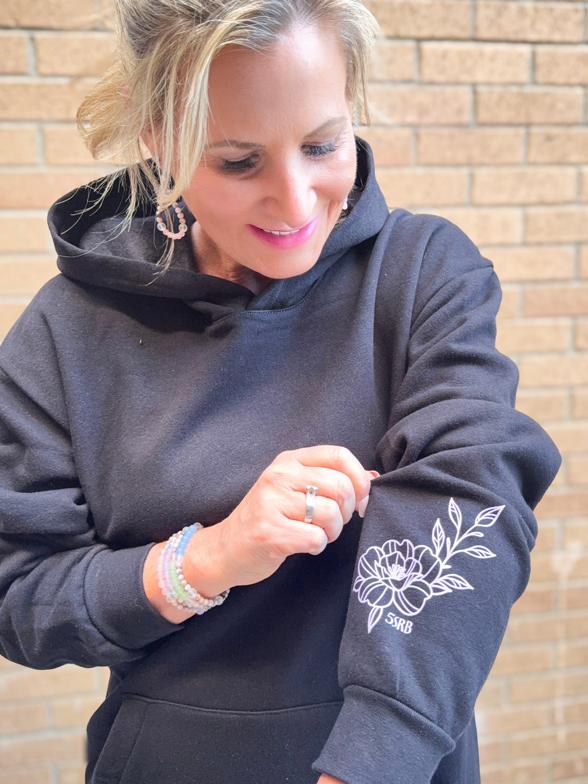 Woman wearing a dark gray hoodie with a floral design on the sleeve, standing against a brick wall.