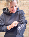 Woman wearing a dark gray hoodie with a floral design on the sleeve, standing against a brick wall.
