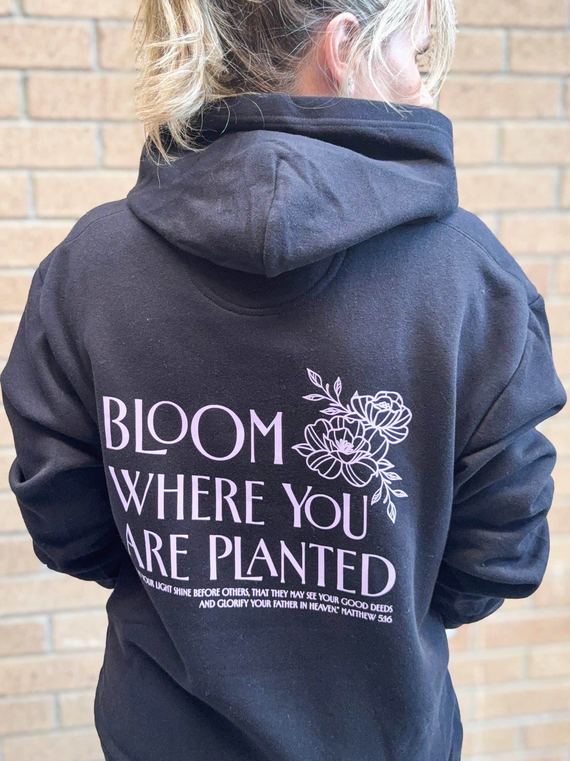 Person wearing a black hoodie with 'Bloom Where You Are Planted' text and floral design on the back, standing against a brick wall.