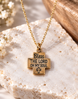 A gold cross pendant necklace with the engraved text 'Bless THE LORD OH MY SOUL' and a small cross at the bottom, displayed on a textured white stone with small white flowers and a wooden bowl in the background.