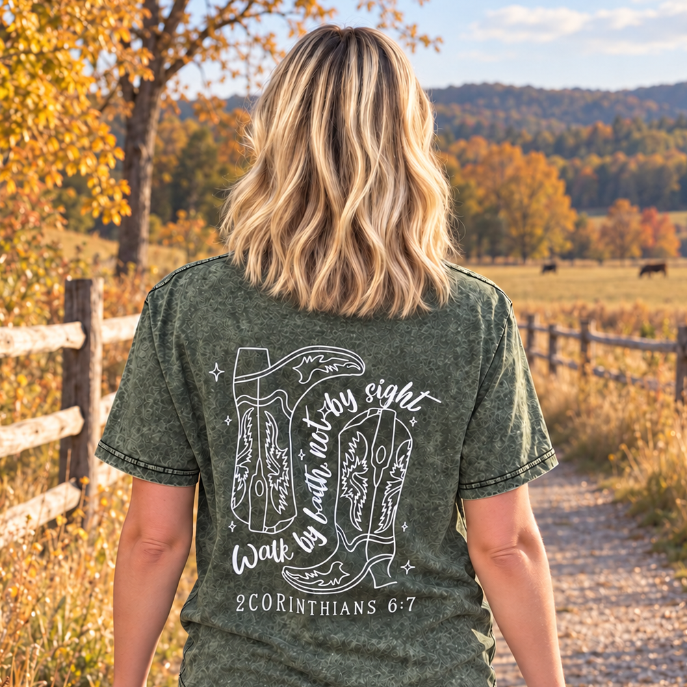 A woman viewed from behind walking on a gravel path in a rural outdoor setting, wearing a dark olive green acid-wash graphic t-shirt with cowboy boots and scripture printed on the back.